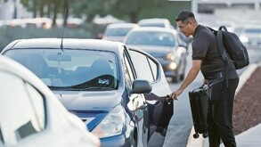Hombre con mochila y maleta abriendo la puerta trasera de un automóvil color azul con otros vehículos transitando en la zona del Aeropuerto de Guadalajara