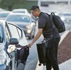 Hombre con mochila y maleta abriendo la puerta trasera de un automóvil color azul con otros vehículos transitando en la zona del Aeropuerto de Guadalajara