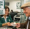 Adulto mayor sonriendo mientras recibe el pago de la pensión del IMSS a través de una ventanilla con una mujer haciendo la entrega de los billetes