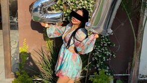 Se observa a una joven con unos globos en la mano, está sonriendo y viste un vestido.