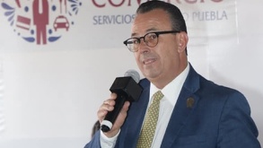 Hombre vestido de traje y usando lentes habla mientras sostiene un micrófono con la mano.