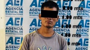 Hombre con los ojos cubiertos junto a una mampara de la Agencia Estatal de Investigación de Oaxaca.