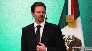 Hombre de traje habla ante un micrófono junto a una bandera de México.