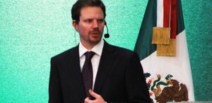 Hombre de traje habla ante un micrófono junto a una bandera de México.