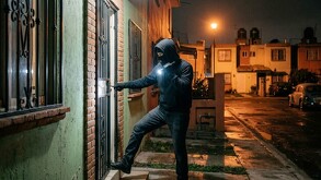 Asaltante con la cara cubierta y vestido de negro apuntando una lámpara para tratar de entrar en una casa durante la noche