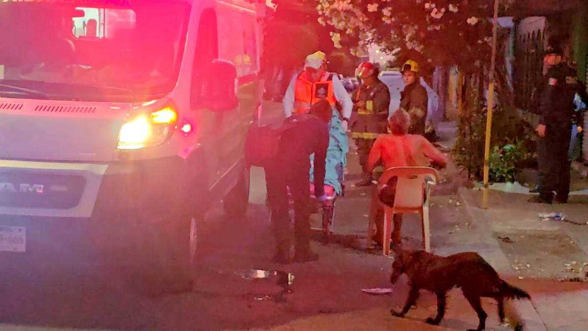 Hombre sin playera sentado en una silla con paramédicos de la Cruz Verde preparando una camilla a un lado de una ambulancia y bomberos alrededor