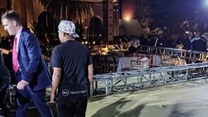 Un hombre de traje visto de perfil caminando y otro visto de espaldas con una estructura metálica caída alrededor de mesas y sillas de una boda en el fondo