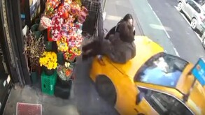 Taxi color amarillo en calles de Nueva York impactando en contra de dos mujeres delante de un puesto con arreglos florales
