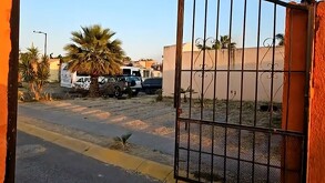 Vista exterior del fraccionamiento Los Agaves con un portón negro abierto y al interior un camión de transporte de personal con dos autos estacionados al ladod
