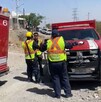Se observa a tres personas con unifromes en tonos amarillos y rojos, y un camión de bomberos rojo.