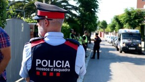 Policía miembro de los Mossos d'Esquadra visto de espaldas volteando hacia la izquierda con otros oficiales y una ambulancia vistos desenfocados en el fondo