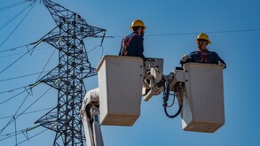 Dos trabajadores de la CFE en canastillas de una grúa realizando trabajos en una torres de luz y cables detrás de ellos