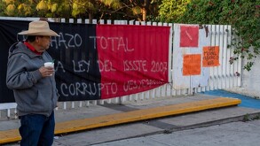 Hombre parado con un vaso de unicel en la mano delante de una reja con barrotes blancos y una bandera rojinegra y carteles de protesta de maestros de la CNTE