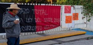 Hombre parado con un vaso de unicel en la mano delante de una reja con barrotes blancos y una bandera rojinegra y carteles de protesta de maestros de la CNTE
