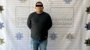 Se observa a un hombre parado, con ojos cubiertos; viste pantalón de mezclilla y playera negra.