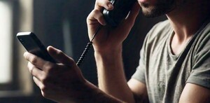 Primer plano enfocado en una mano sosteniendo un celular, con la silueta borrosa de una persona con capucha oscura al fondo.