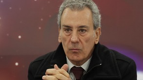 Hombre canoso vistiendo una corbata cruza las manos y mira hacia el frente.