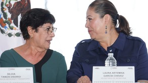 Dos mujeres mayores hablando de frente con una bandera de México al fondo.