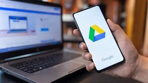 Persona sosteniendo un teléfono celular con la app de Google Drive abierta y con una laptop desenfocada en el fondo