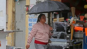 Mujer caminando delante de una tienda de diversos utensilios con una sombrilla negra ligeramente mojada y vistiendo una chamarra color rosa