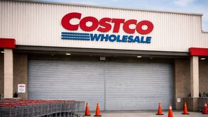 Entrada de una tienda Costco con la cortina metálica abajo, carritos alineados al costado y conos naranjas frente al acceso principal.