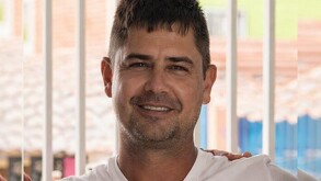 Eduardo Ochoa Arias viendo de frente a la cámara mientras sonríe y viste una playera blanca con barrotes desenfocados en el fondo