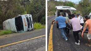 una urvan volcada a un lado de un camino y personas trasladando a un pasajero lesionado