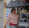 Mujer caminando delante de una tienda de diversos utensilios con una sombrilla negra ligeramente mojada y vistiendo una chamarra color rosa