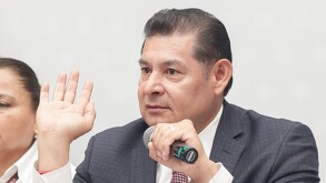 Hombre de traje sostiene un micrófono mientras hace un ademán con una de sus manos.