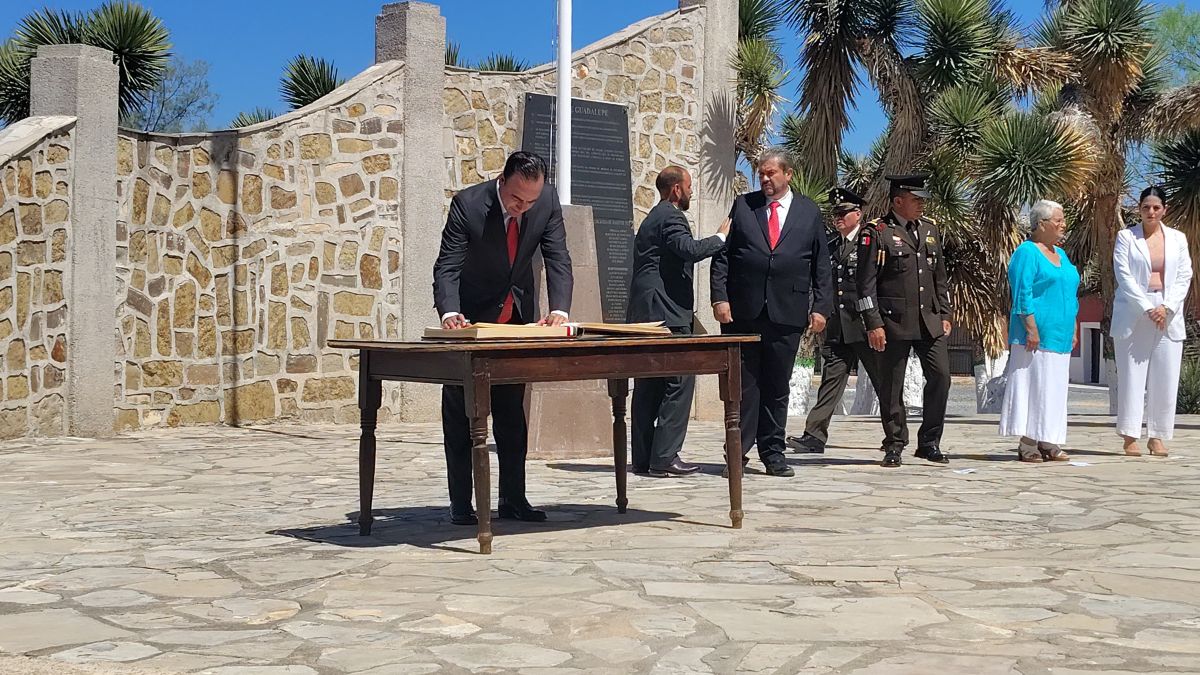 Acto conmemorativo al aire libre donde autoridades y asistentes se reúnen en un espacio histórico; parte de la imagen se encuentra cubierta por un objeto en pri