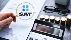 Mano sosteniendo una lupa con el logo del SAT delante de un documento con una calculadora, monedas y unos lentes a un lado