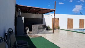 Patio amplio con piso de loseta, área techada tipo palapa con asador, barra y refrigerador, sillas apiladas a un costado y una alberca al fondo, bajo un cielo d