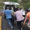 una urvan volcada a un lado de un camino y personas trasladando a un pasajero lesionado