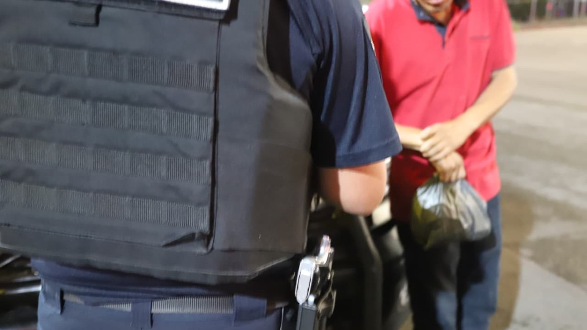 Policía de Tlaquepaque con chaleco antibalas y una pistola delante de un hombre con playera roja sosteniendo una bolsa