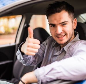 Un joven conductor sonríe mientras está al volante de su automóvil y hace una señal de aprobación con el pulgar.