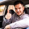 Un joven conductor sonríe mientras está al volante de su automóvil y hace una señal de aprobación con el pulgar.