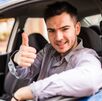 Un joven conductor sonríe mientras está al volante de su automóvil y hace una señal de aprobación con el pulgar.
