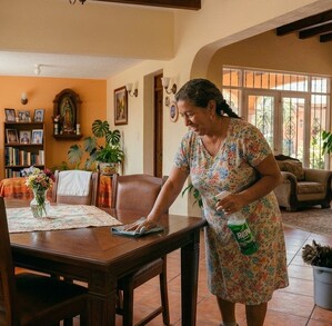 Se observa a una mujer limpiando una mesa de madera, está dentro de una casa.