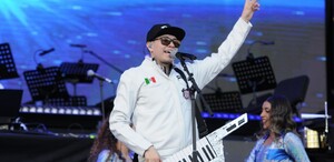 Hombre con un teclado cantando, mientras apunta con la mano hacia el cielo.