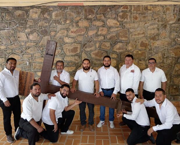 Un grupo de hombres vestidos con camisas blancas posan en conjunto mientras sostienen una gran cruz de madera en un espacio con piso de loseta y una pared de pi