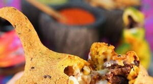 Una persona sostiene una quesadilla dorada con forma creativa, abierta para mostrar su relleno de carne y queso, con limones y salsas desenfocados al fondo.