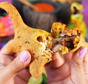 Una persona sostiene una quesadilla dorada con forma creativa, abierta para mostrar su relleno de carne y queso, con limones y salsas desenfocados al fondo.