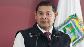 Hombre parado delante de una bandera del estado de Puebla.