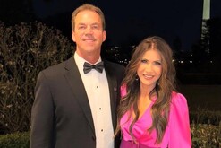 Kristi Noem vistiendo un vestido rosa junto a su esposo Bryon Noem vestido de traje delante de un jardín