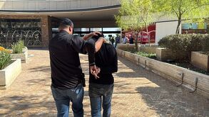 Un hombre es detenido por otra persona en un espacio público al exterior de un edificio comercial; el detenido tiene las manos sujetas detrás de la espalda mien