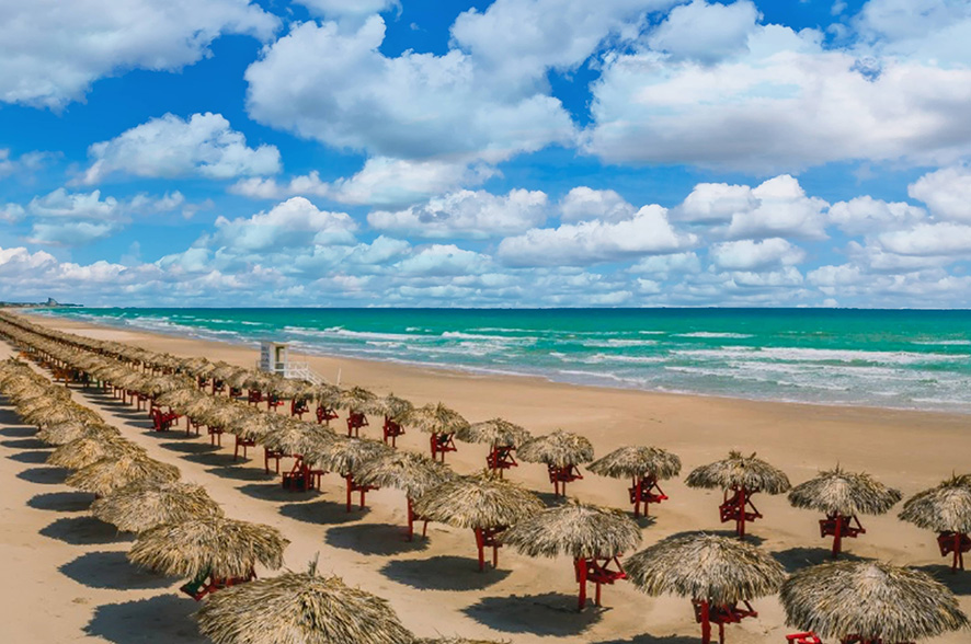 Vista panorámica de Playa Miramar con filas de palapas sobre la arena, el mar en tonos turquesa y un cielo parcialmente nublado que acompaña el paisaje costero.