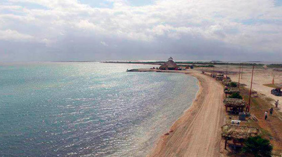 Vista de Playa La Carbonera con una franja de arena que bordea el agua tranquila, palapas dispersas a lo largo de la orilla y un cielo mayormente nublado que cu