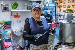 Mujer dentro de la cocina de un comercio en Jalisco sosteniendo unos cuchillos con ollas al frente y personas en el fondo