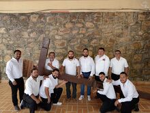 Un grupo de hombres vestidos con camisas blancas posan en conjunto mientras sostienen una gran cruz de madera en un espacio con piso de loseta y una pared de pi