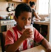 Se observa a un niño moreno que está tomando un vaso de leche color rosa, está sentado en una mesa.
